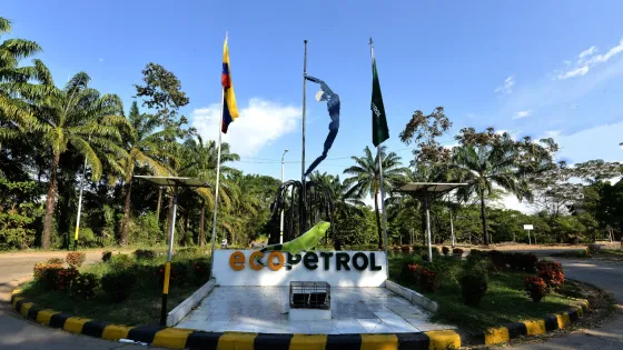 Entrada principal de una planta de Ecopetrol rodeada de palmeras, con las banderas de Colombia y de la empresa ondeando junto a una escultura metálica y el logotipo de Ecopetrol en primer plano.